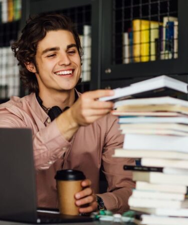 Estudante sorridente segura livros ao lado de um notebook durante os estudos.