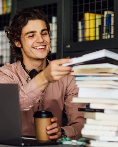 Estudante sorridente segura livros ao lado de um notebook durante os estudos.