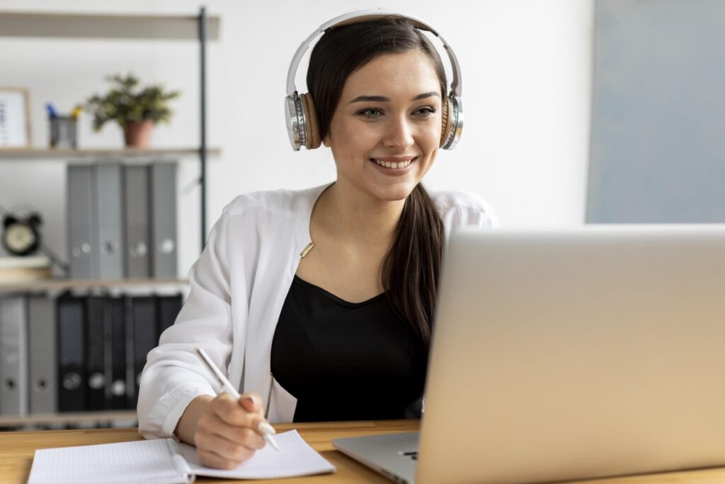 Mulher usando fones de ouvido e fazendo anotações enquanto estuda no computador, representando a rotina de quem busca uma pós-graduação em Educação.