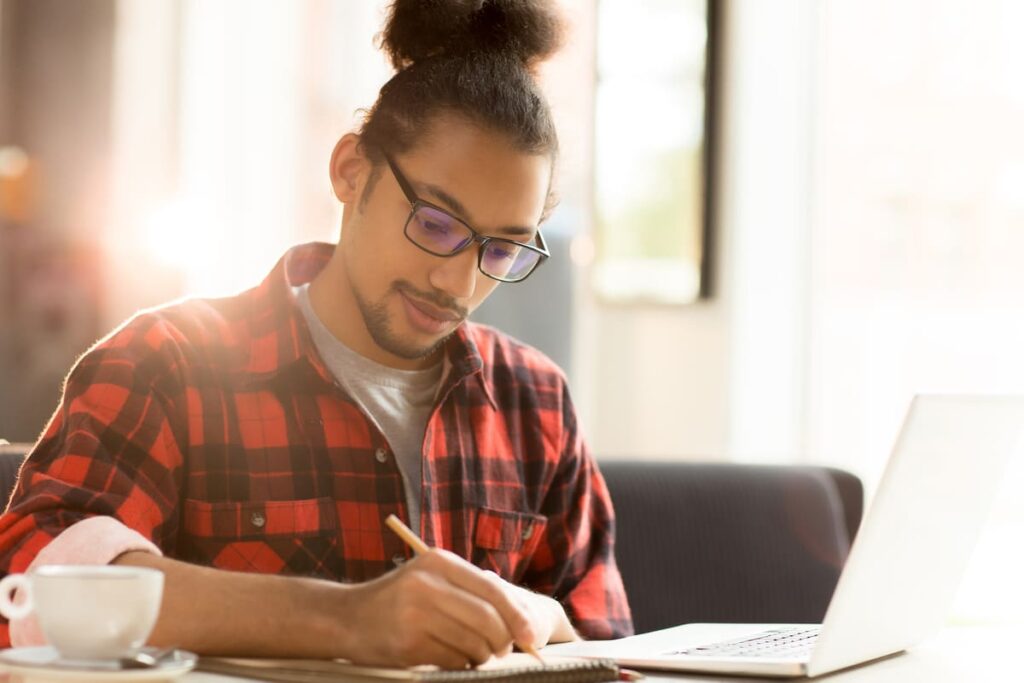 Estudante usando óculos estuda e faz anotações ao lado de um notebook.