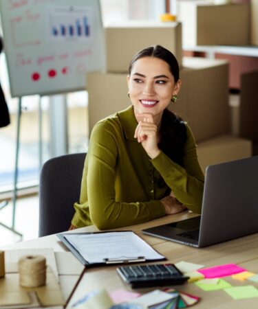 Mulher jovem olha para cima sorridente com a mão no queixo. Ao seu redo, existem elementos que remetem a moda, como roupas em cabideiros e manequim. Em cima de sua mesa, estão alguns post its, calculadora e uma prancheta.