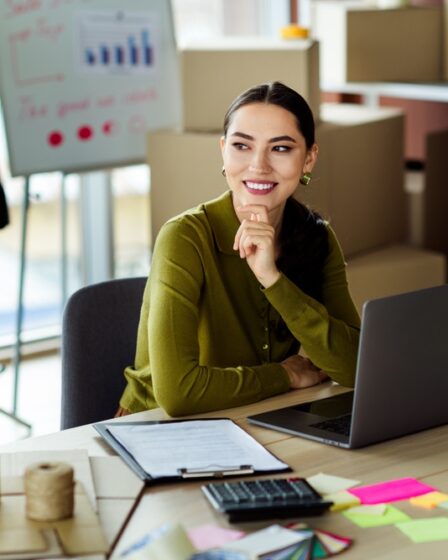 Mulher jovem olha para cima sorridente com a mão no queixo. Ao seu redo, existem elementos que remetem a moda, como roupas em cabideiros e manequim. Em cima de sua mesa, estão alguns post its, calculadora e uma prancheta.