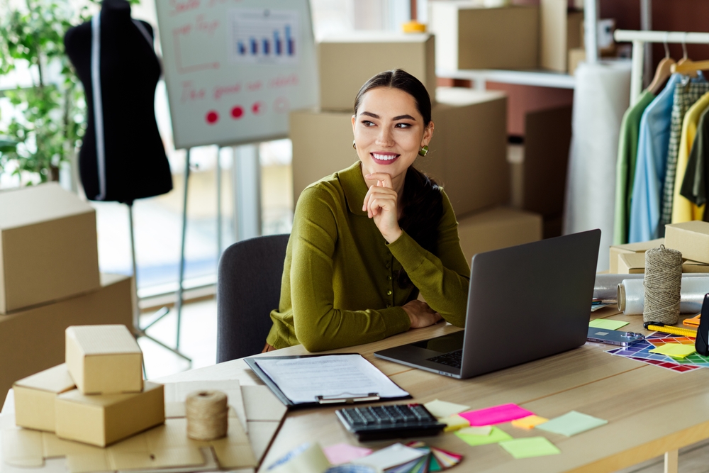 Mulher jovem olha para cima sorridente com a mão no queixo. Ao seu redo, existem elementos que remetem a moda, como roupas em cabideiros e manequim. Em cima de sua mesa, estão alguns post its, calculadora e uma prancheta.