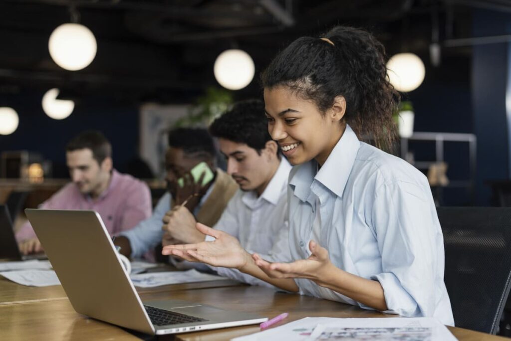 Profissional em ambiente de trabalho usando notebook com colegas ao fundo, mostrando que quem faz pós-graduação pode estagiar.