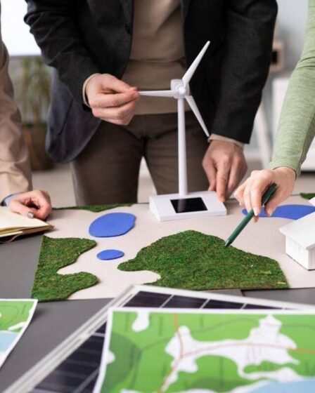 Equipe de profissionais planeja soluções de energia renovável sobre uma maquete com turbina eólica, ilustrando projetos sustentáveis e a formação da pós em Engenharia Ambiental.